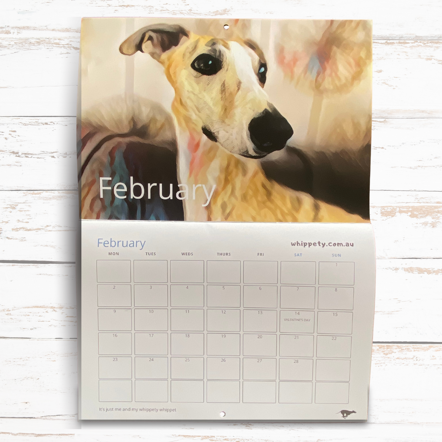 Wall calendar for February with a whippet dog on the cover, displayed on a wooden surface.